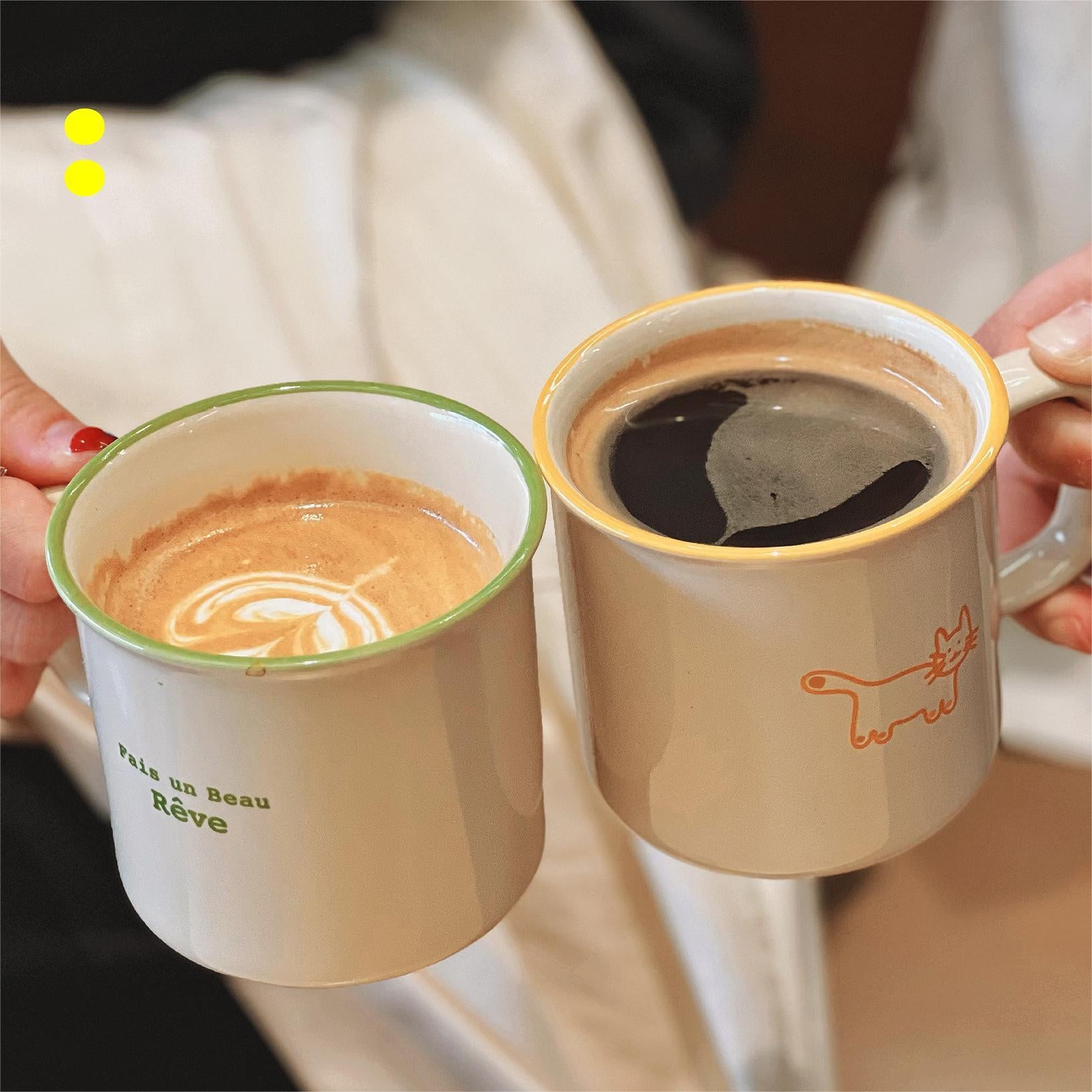 Friends holding the Fais un Beau Rêve Coffee Mugs Cheers. One hold the Matcha Colors and Another holds the Orange. Latte and Americano in Mugs.